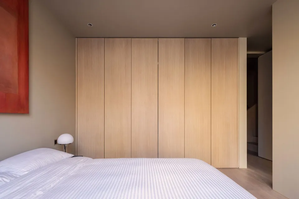 A minimalist bedroom featuring a neatly made bed with a striped white cover, a modern lampshade, and a wooden paneled wall.