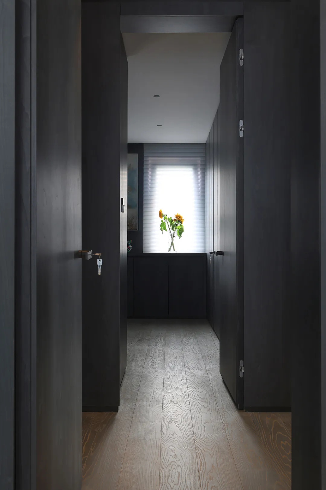 A modern, minimalist corridor with black walls and a wooden floor, showcasing sleek doors and a window with sunflowers.