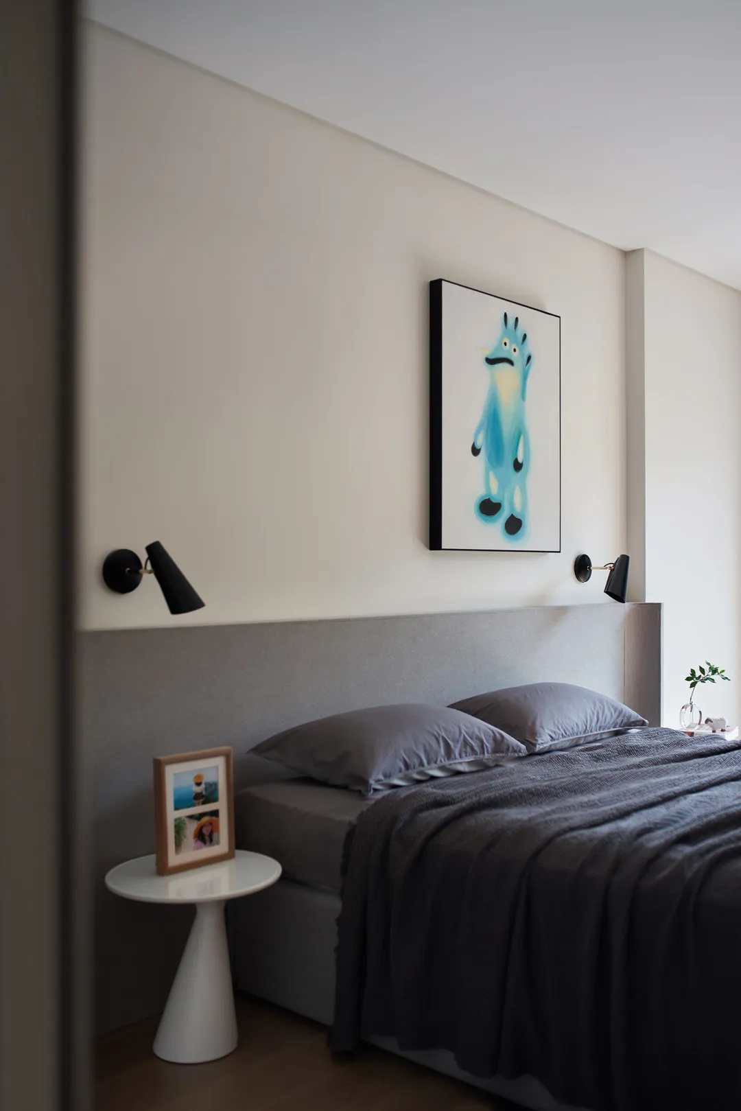 A minimalist bedroom with a grey fabric bed and white side table.