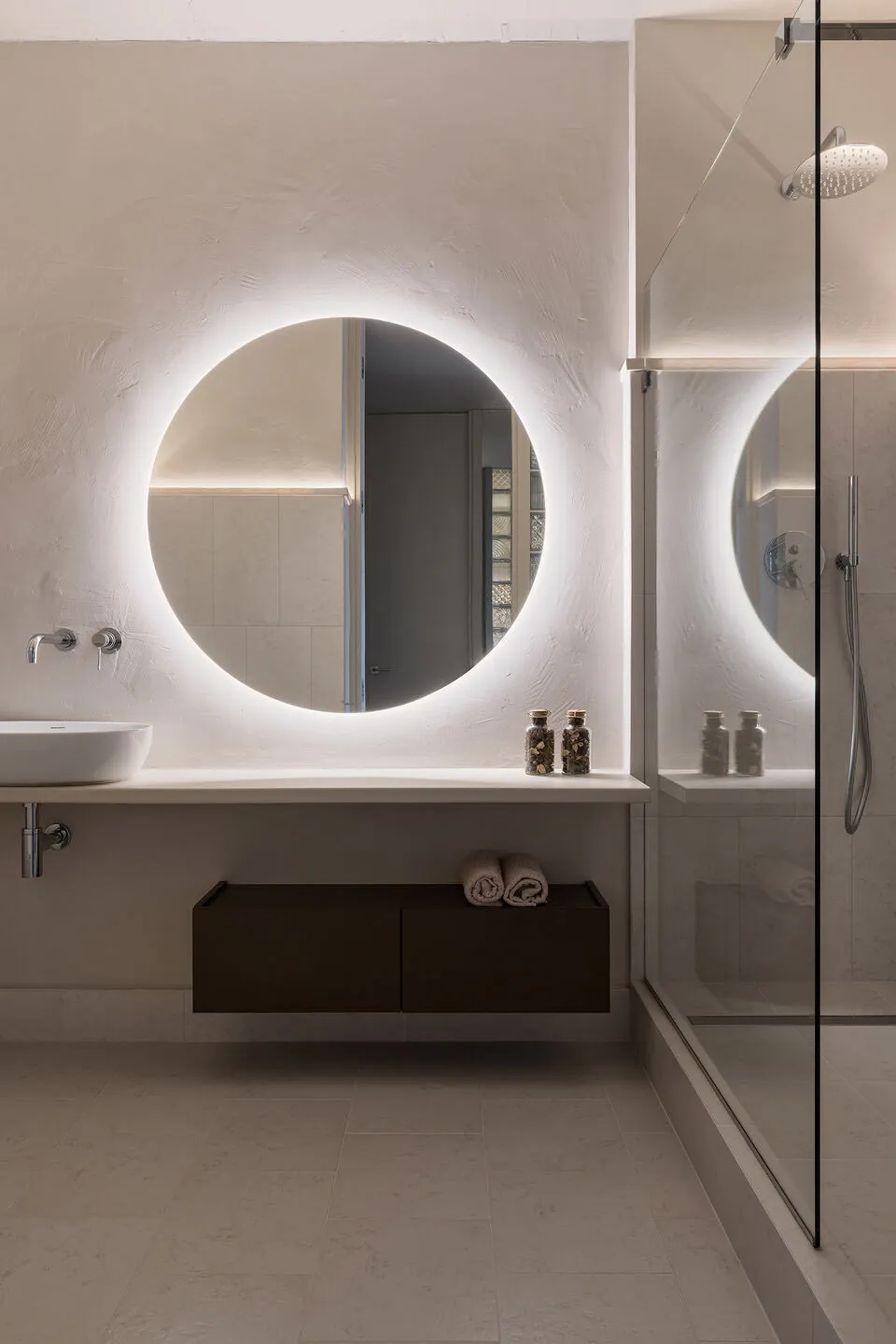 Modern minimalist bathroom with circular mirror featuring backlighting and concrete floor and walls, providing a sleek and co