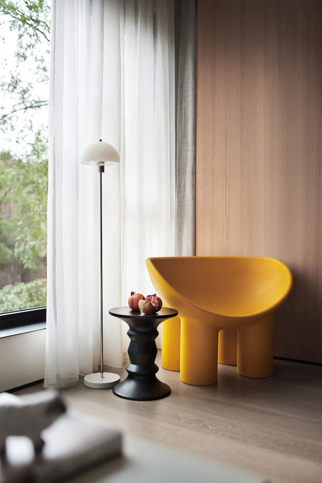 A modern living room featuring a yellow plastic chair, a black side table, and a floor lamp near a window with sheer curtains