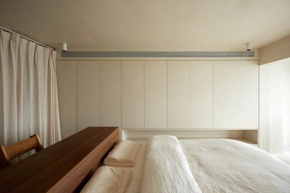 A minimalist bedroom with a textured wooden bed frame and neutral-colored bedding against a white wall.