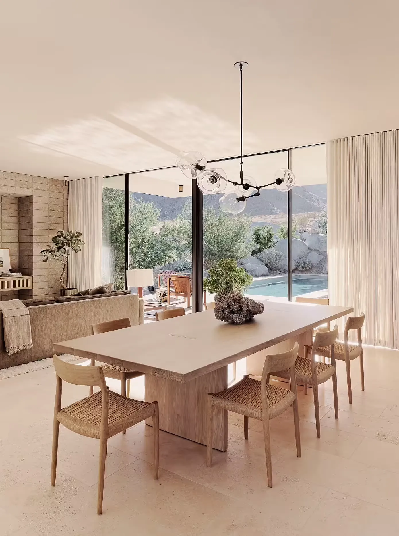 Modern dining room featuring a large natural wood dining table with six chairs, a minimalist chandelier, and a plush couch wi