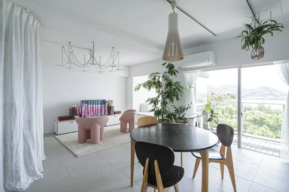 A modern living and dining room featuring a round wooden table, pink sculptural chairs, and large indoor plants.