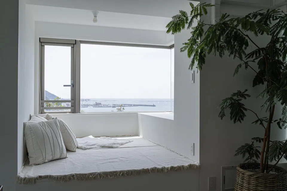 White minimalistic window seat overlooking the sea, with green leafy houseplant and light fixtures.