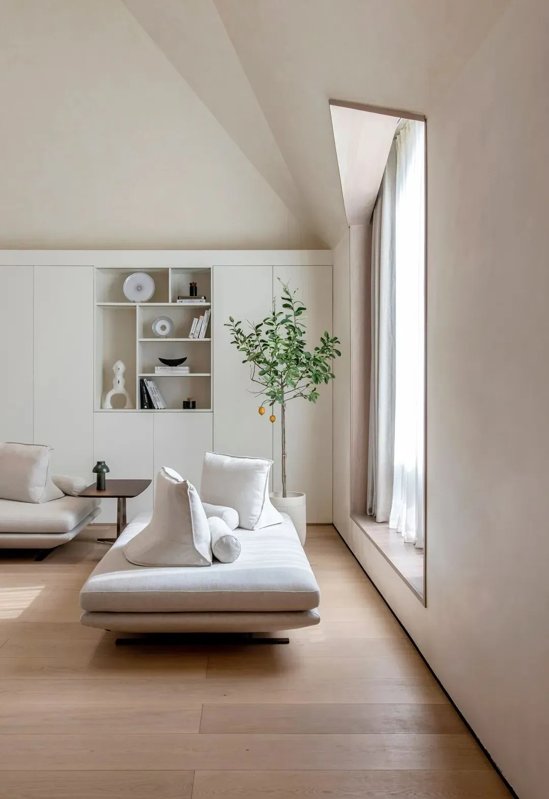 A minimalistic residential interior with neutral tones featuring a daybed, bookshelf, and window