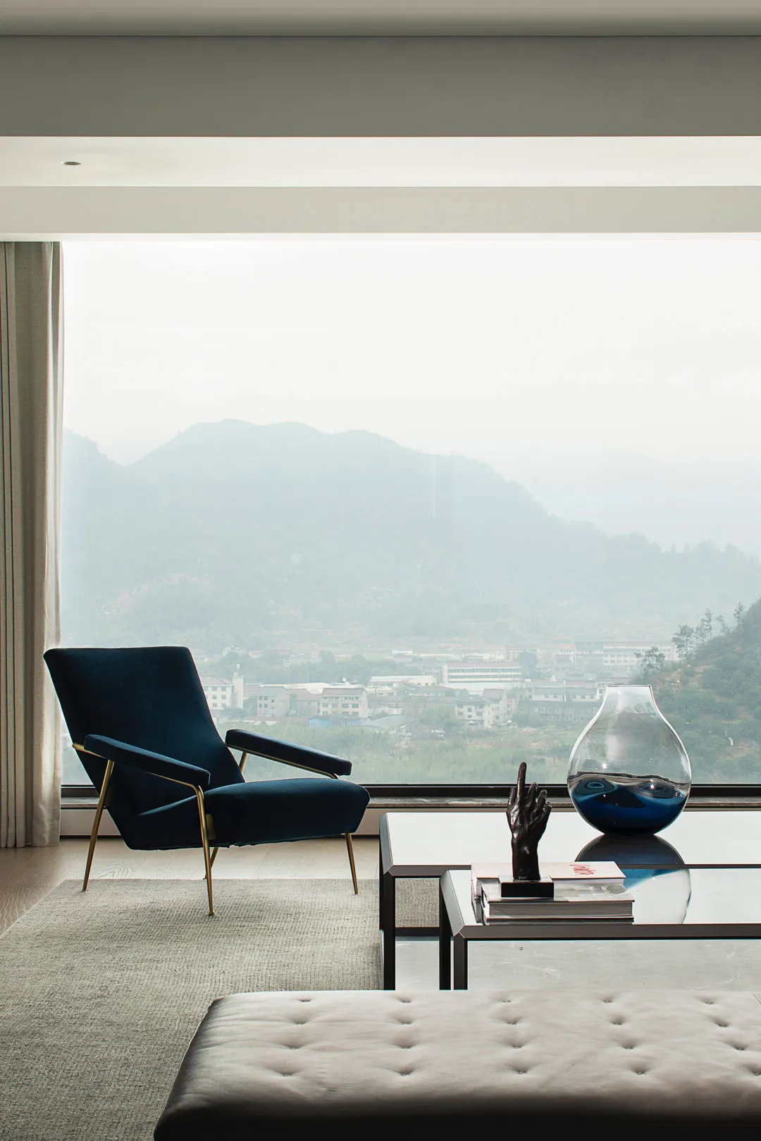 Modern living room featuring a glass coffee table, plush carpet, and a velvet armchair by a large window.