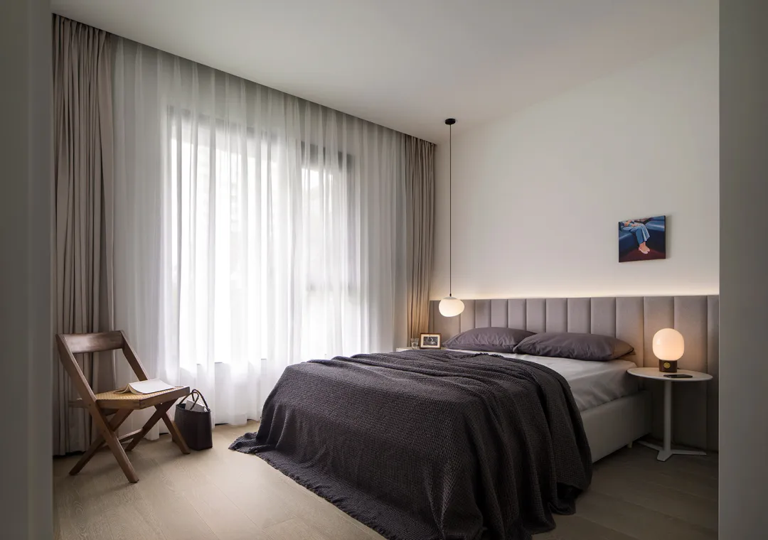 A minimalist bedroom with a gray fabric bed and white curtains, featuring a modern design with soft lighting and a simple col