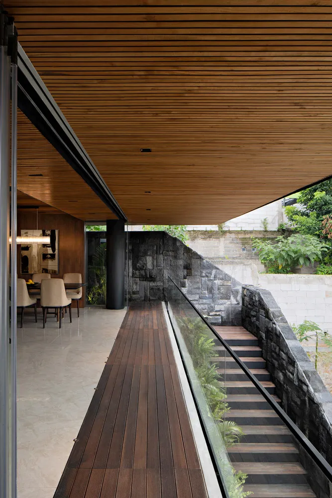 Modern residential exterior with wooden deck, stone wall, and glass railings.