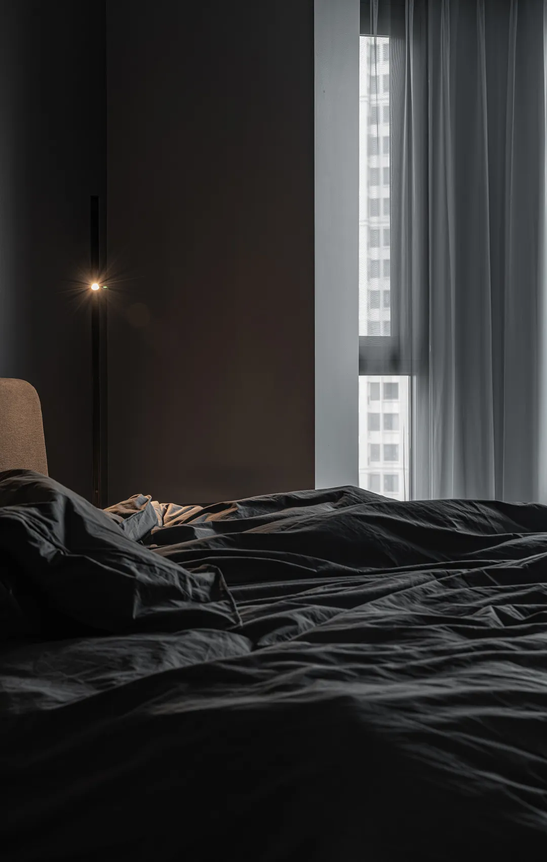 A minimalist bedroom with a black bed and fabric bedding, positioned near a window with sheer curtains and an adjacent floor