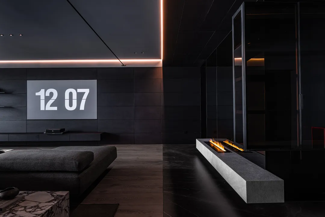 A modern minimalist living space featuring a concrete fireplace and black walls with a clock displaying the time 12:07.