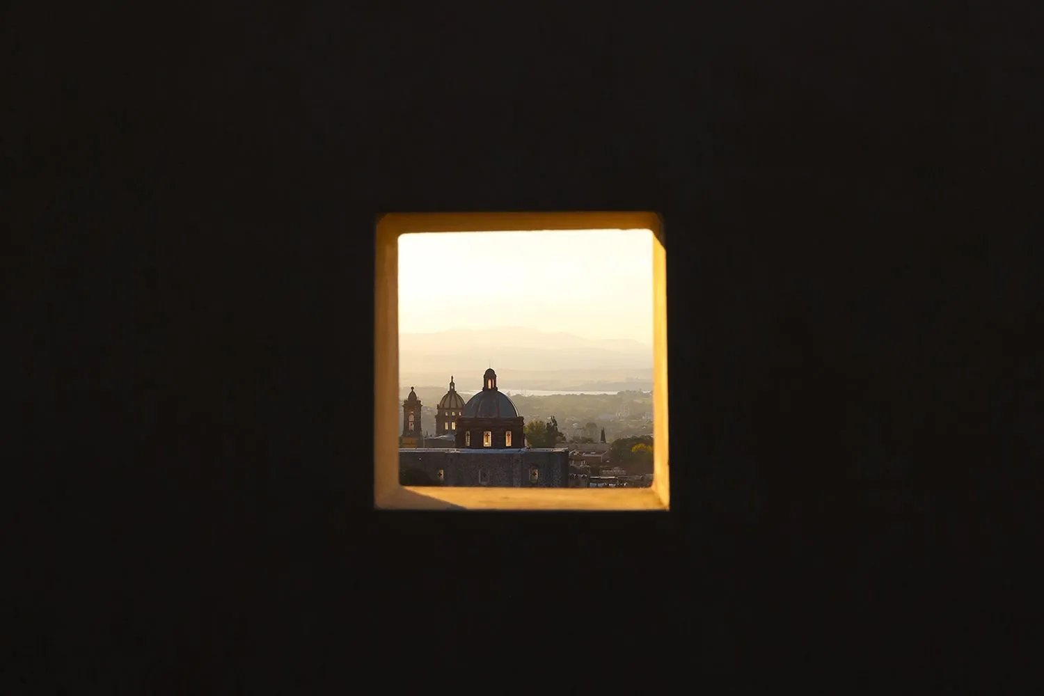 A square window frame reveals a view of a cityscape with domed structures and mountains in the distance, illuminated by a gol