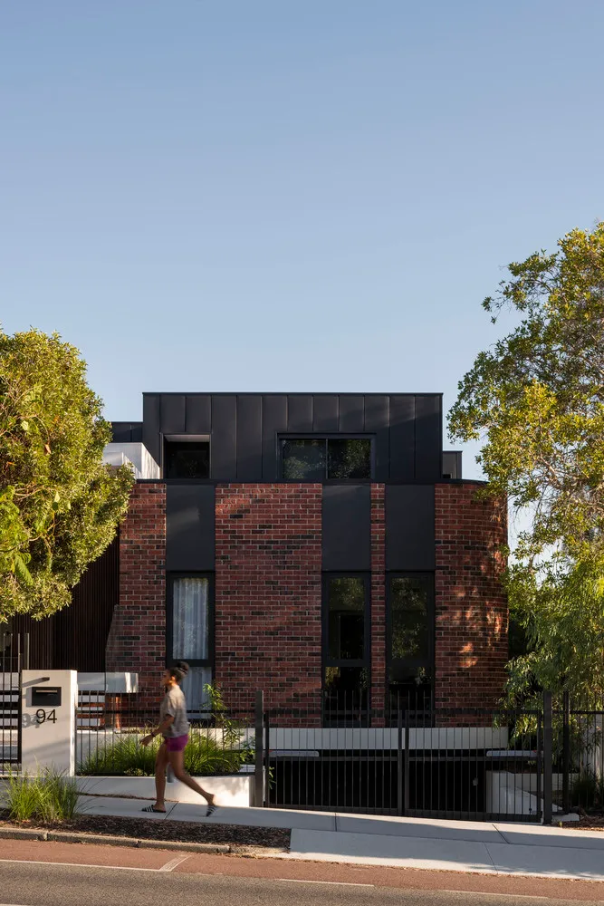A modern residential house with a brick facade and black accents, set against a clear blue sky with lush green trees surround