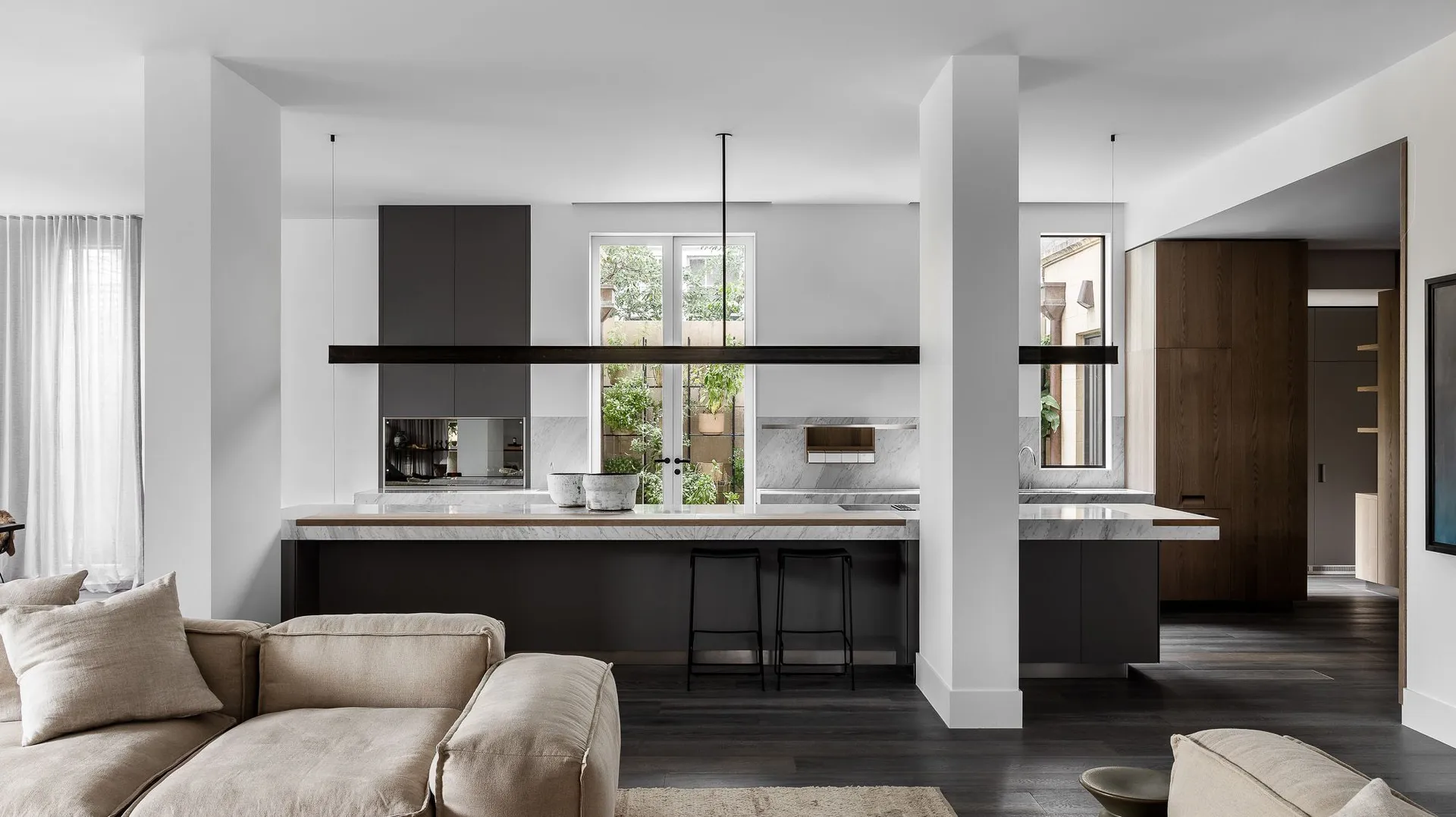 Modern kitchen with marble countertops and wooden cabinets, featuring metal stools and a view of the outdoor garden.