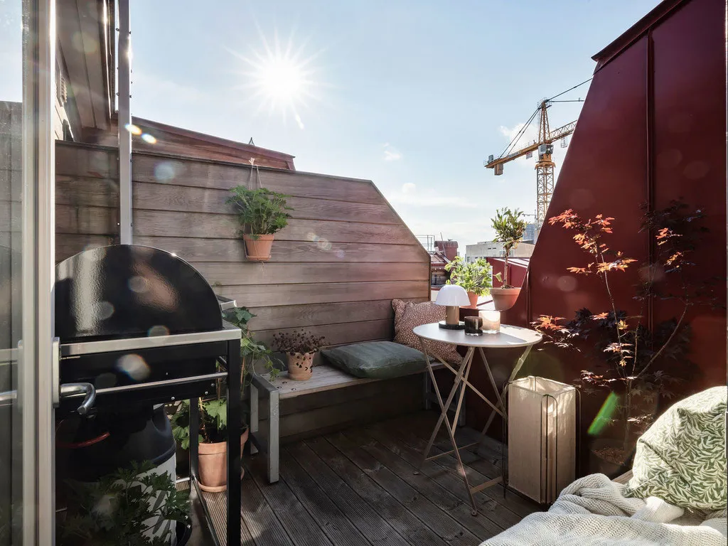 Urban rooftop space featuring a wooden bench, metal grill, and potted plants.