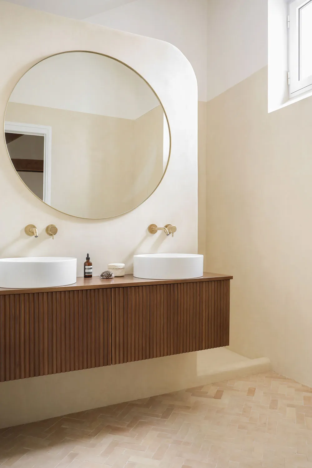 A modern bathroom features a wooden vanity with ceramic sinks and a large circular mirror.