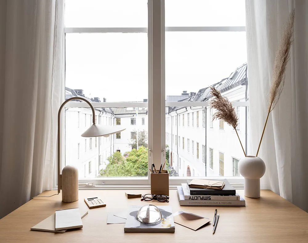 A modern desk setup featuring a white vase, lamp, and books against a window with a view of buildings.