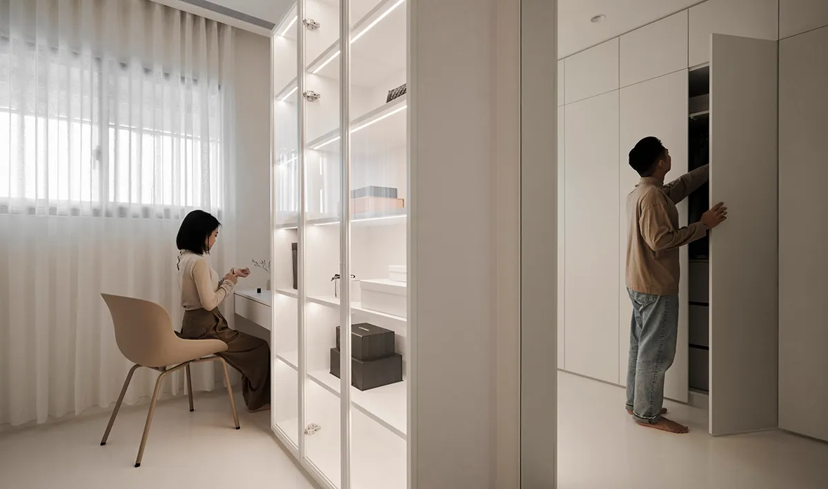 A modern minimalist apartment with a woman sitting on a chair and a man standing in front of a wardrobe.