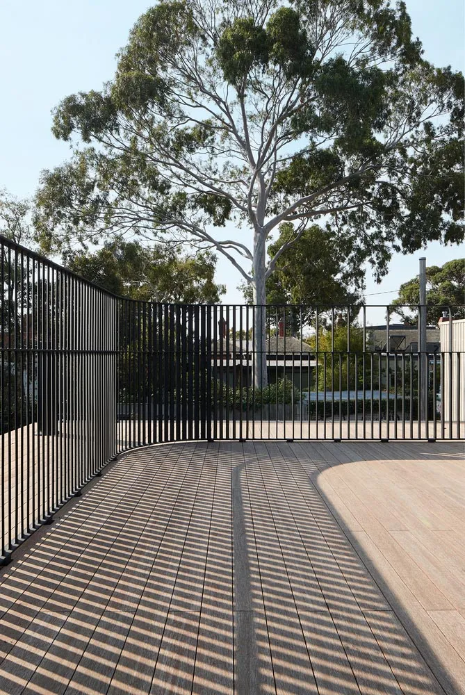 A modern minimalist outdoor patio with wooden flooring and black metal railings, surrounded by trees and natural light.