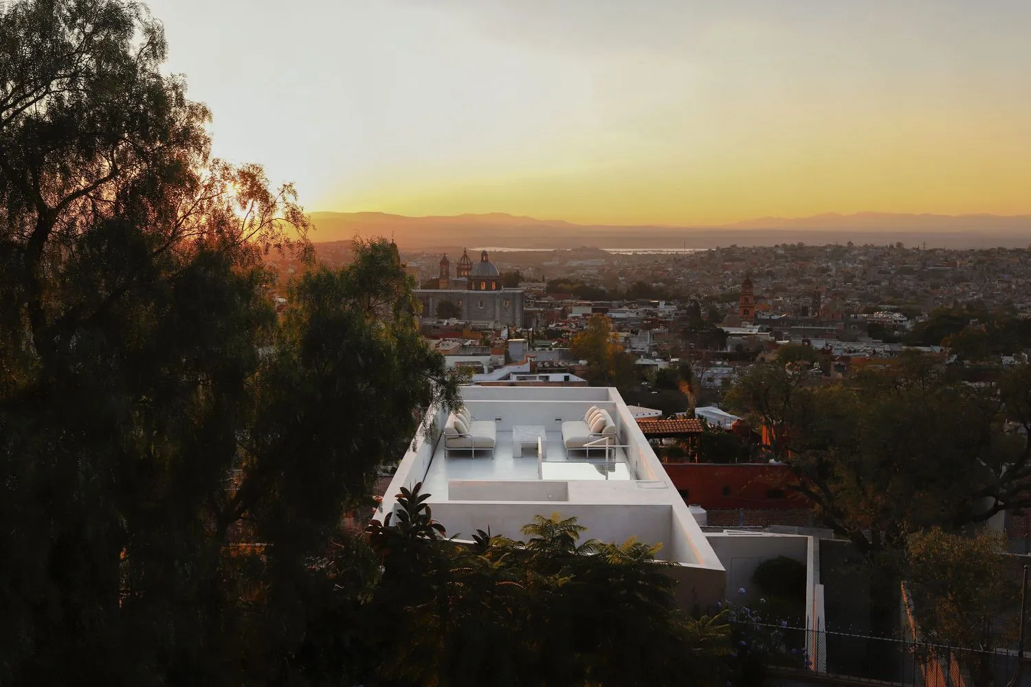 A rooftop with white furniture overlooking a city under a sunset sky.