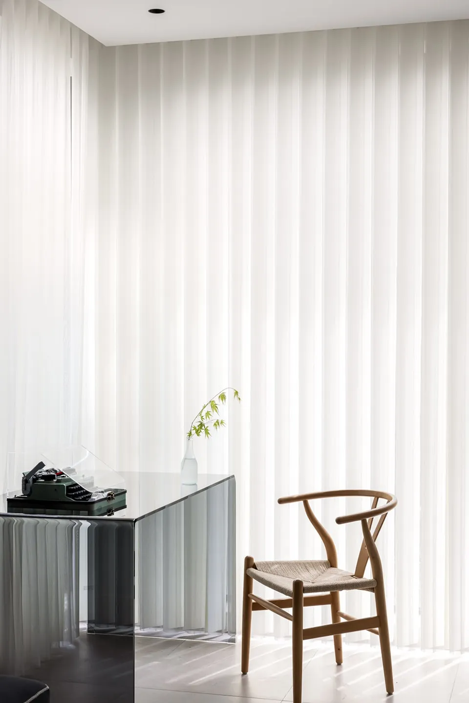 A minimalist desk space featuring a glass desk and a wooden chair with a single stem of greenery.