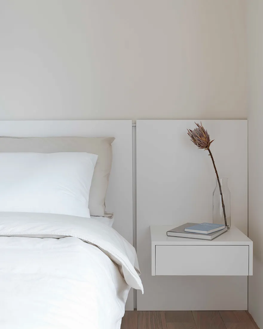 A serene, minimalist bedroom featuring a white bed with neatly arranged white bedding and a simple, white nightstand with a v