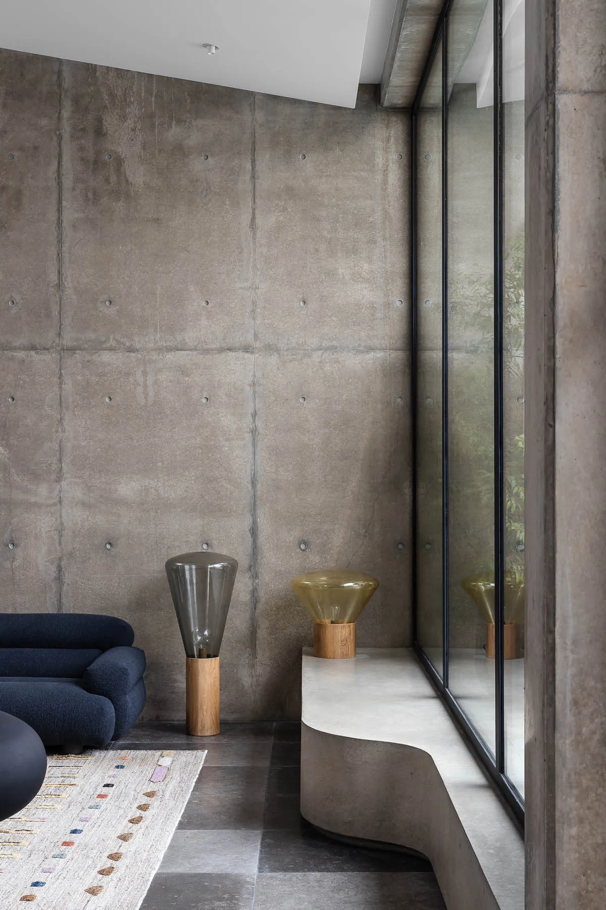 A modern minimalist living room featuring a dark blue sofa, a curved concrete bench, and geometric floor lamp on a patterned
