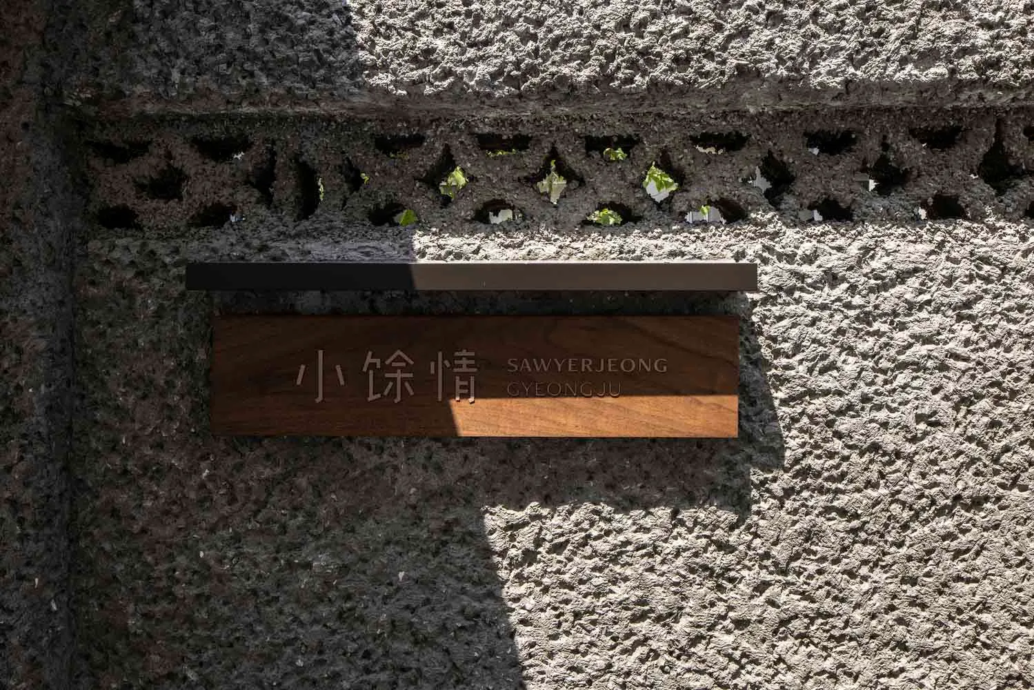 A wooden sign engraved with Chinese characters and English text, mounted on a concrete storefront wall with decorative perfor