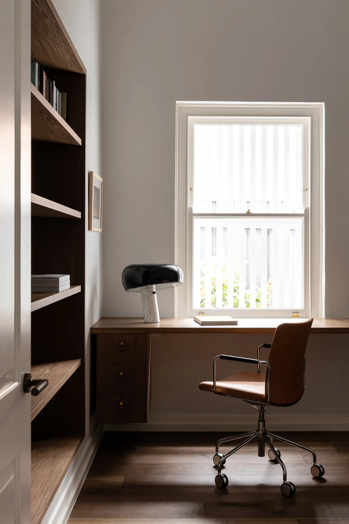 A modern home office with a desk, chair, and lamp next to a bookshelf.