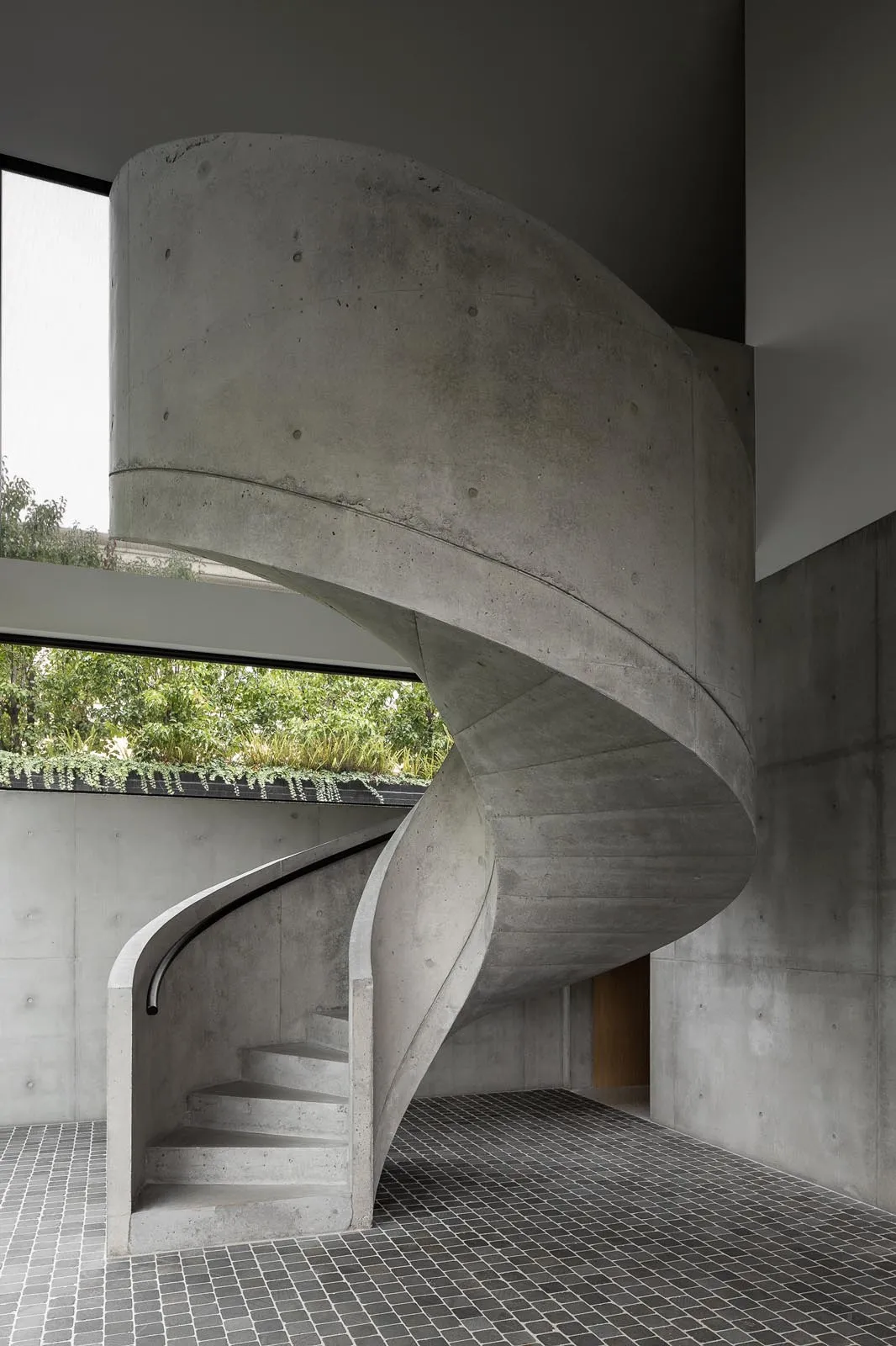 Circular concrete staircase with a dark rail in an industrial space with large windows.