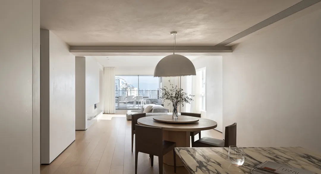 A minimalist dining room with a round wooden table and chairs, accented by a modern hanging light fixture and a vase with flo
