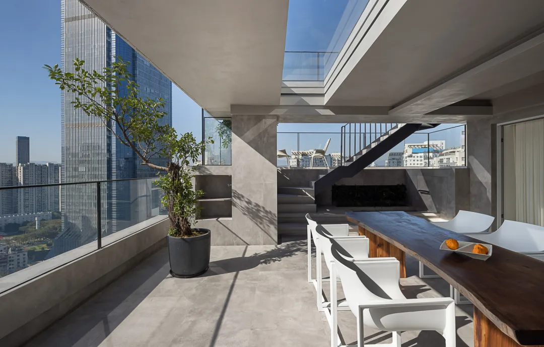 A minimalist balcony design featuring concrete surfaces, white chairs, and a wooden table with oranges.