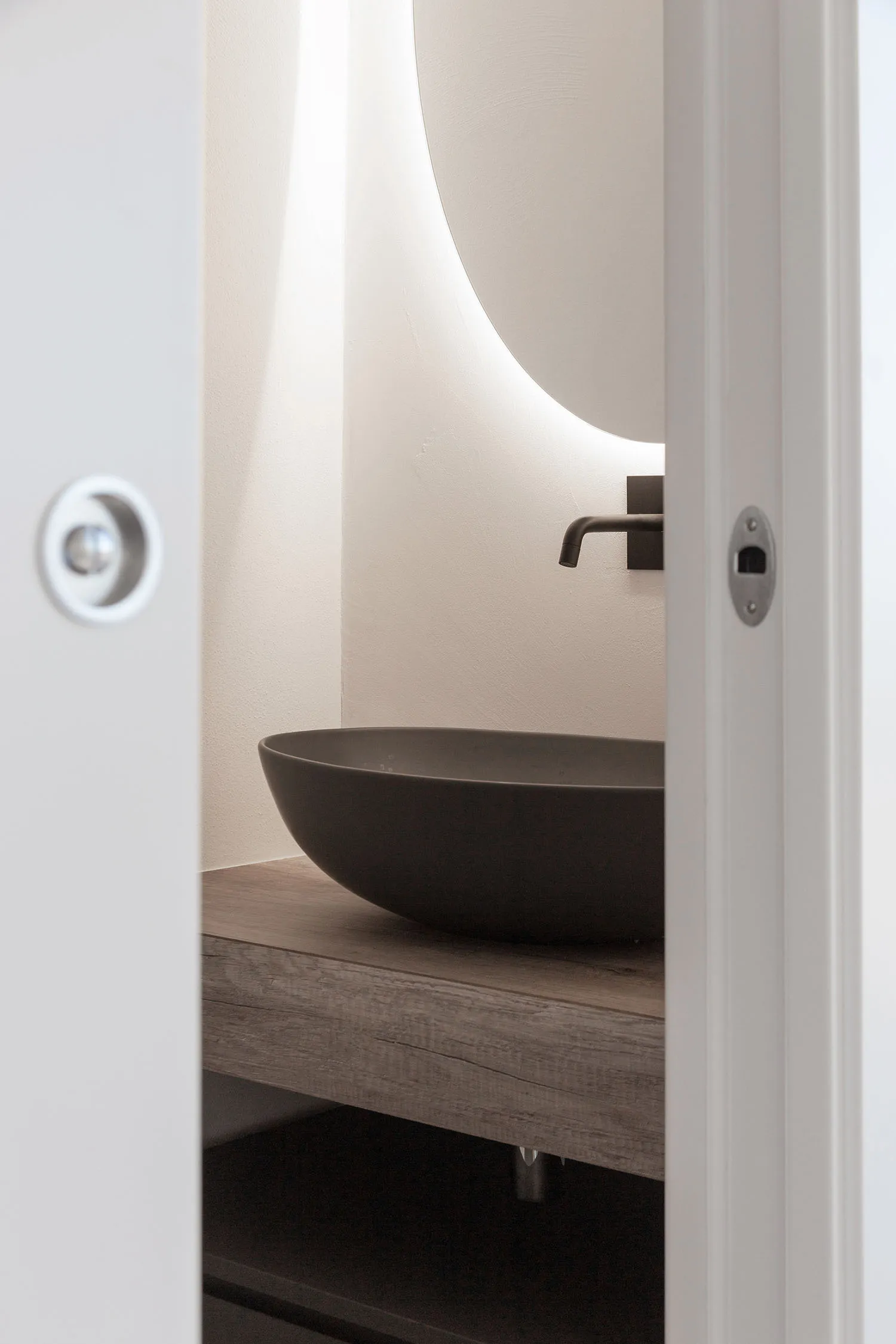 Modern bathroom featuring a sleek, dark-colored concrete sink and illuminated oval-shaped mirror above a floating wooden coun