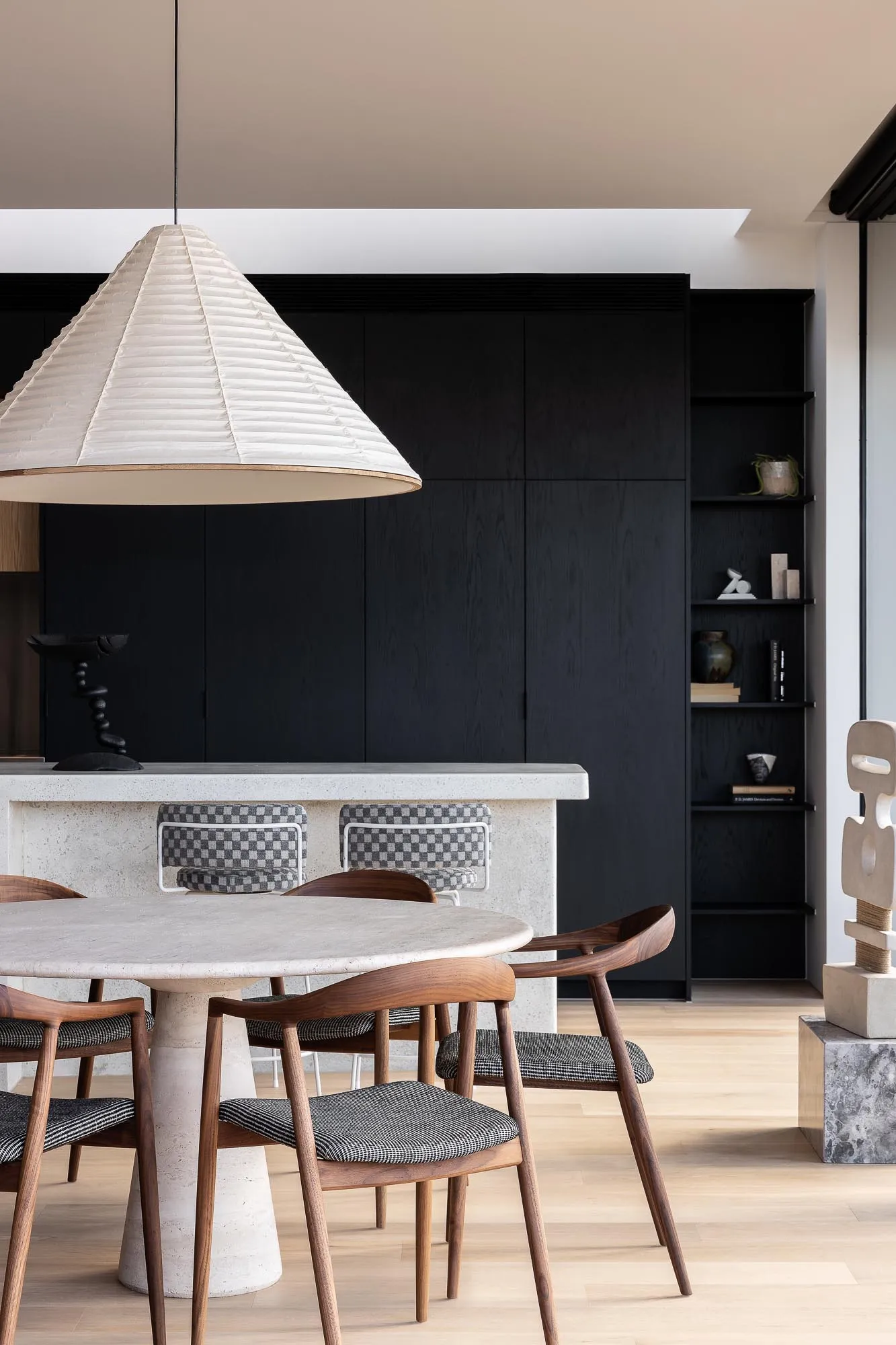 A modern dining room featuring a concrete table and chairs, with a large lampshade overhead and a black shelving unit in the