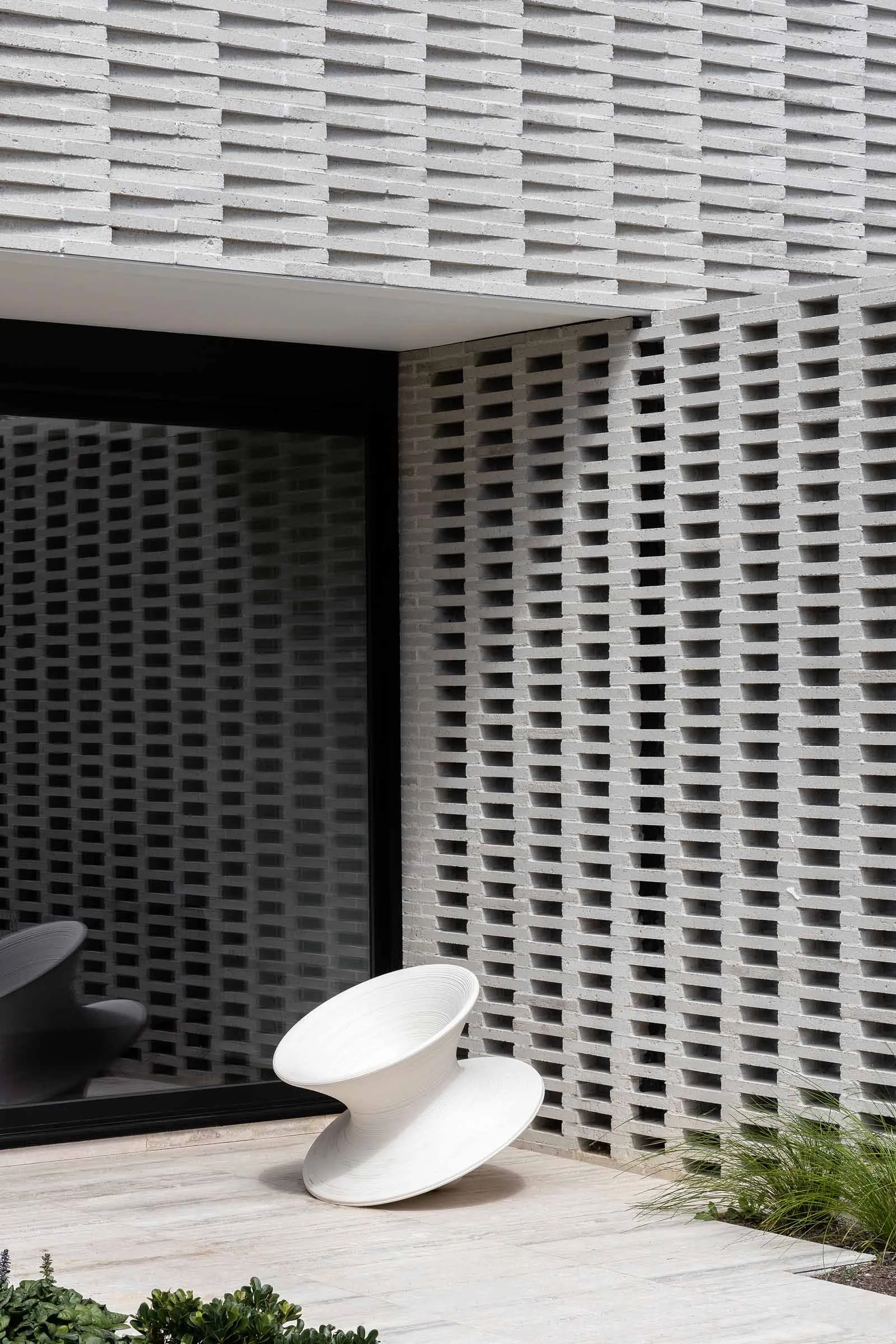 A modern exterior design featuring a concrete wall with a grid pattern and a unique, angular white chair in front of a large