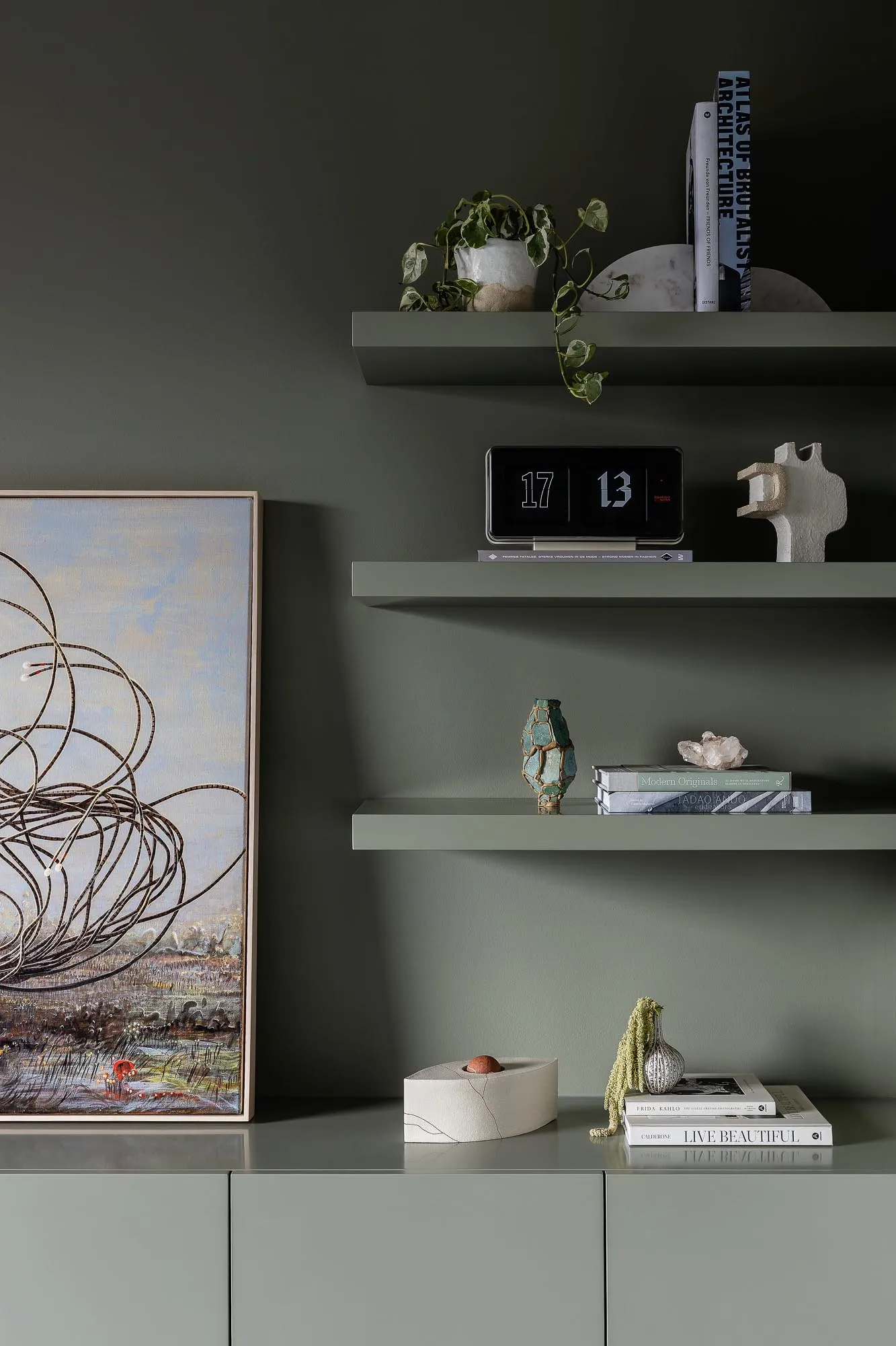 Modern floating shelves with green accents featuring vases, a clock, books, and plants against a dark green wall.