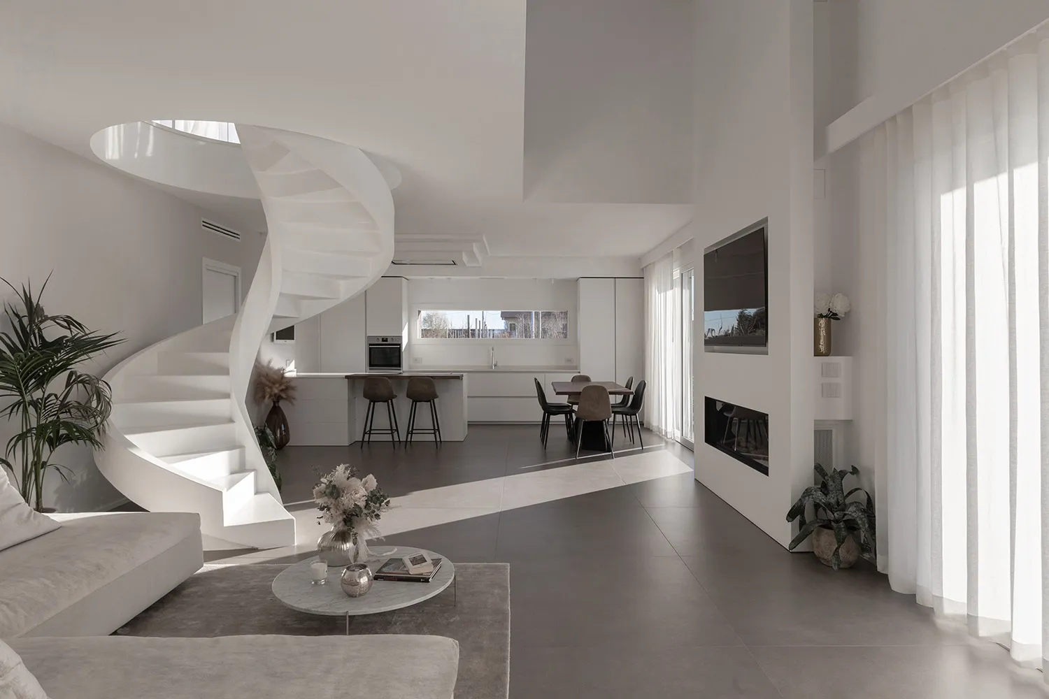 A modern living room featuring a white stone floor, a spiral staircase, a round coffee table, a minimalist sofa, and a kitche