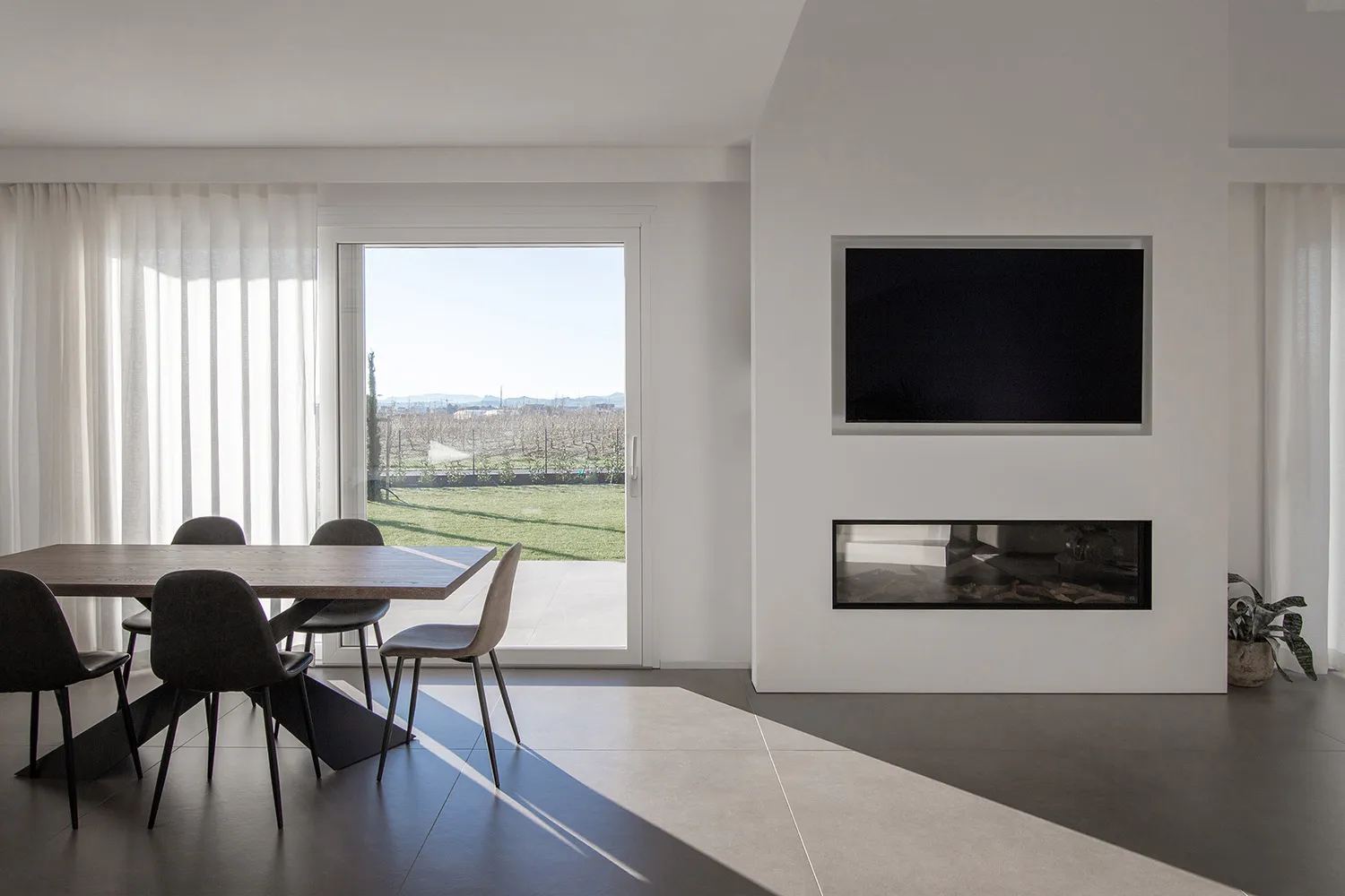 A modern minimalist living room with a concrete floor, a fireplace, a television, and a dining area with a wooden table and c