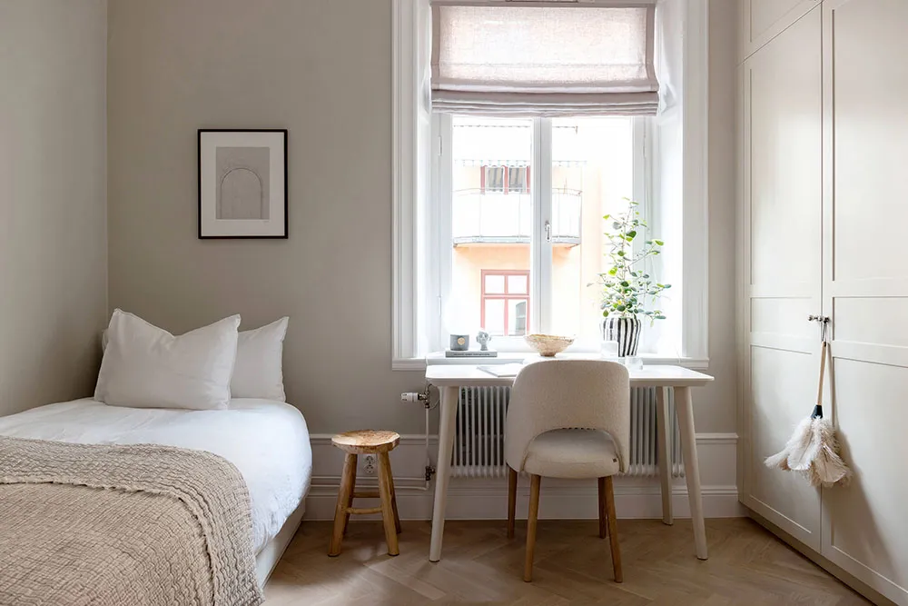 Bedroom filled with light, neutral tones, featuring a bed, a desk, a stool, and a window with a plant.