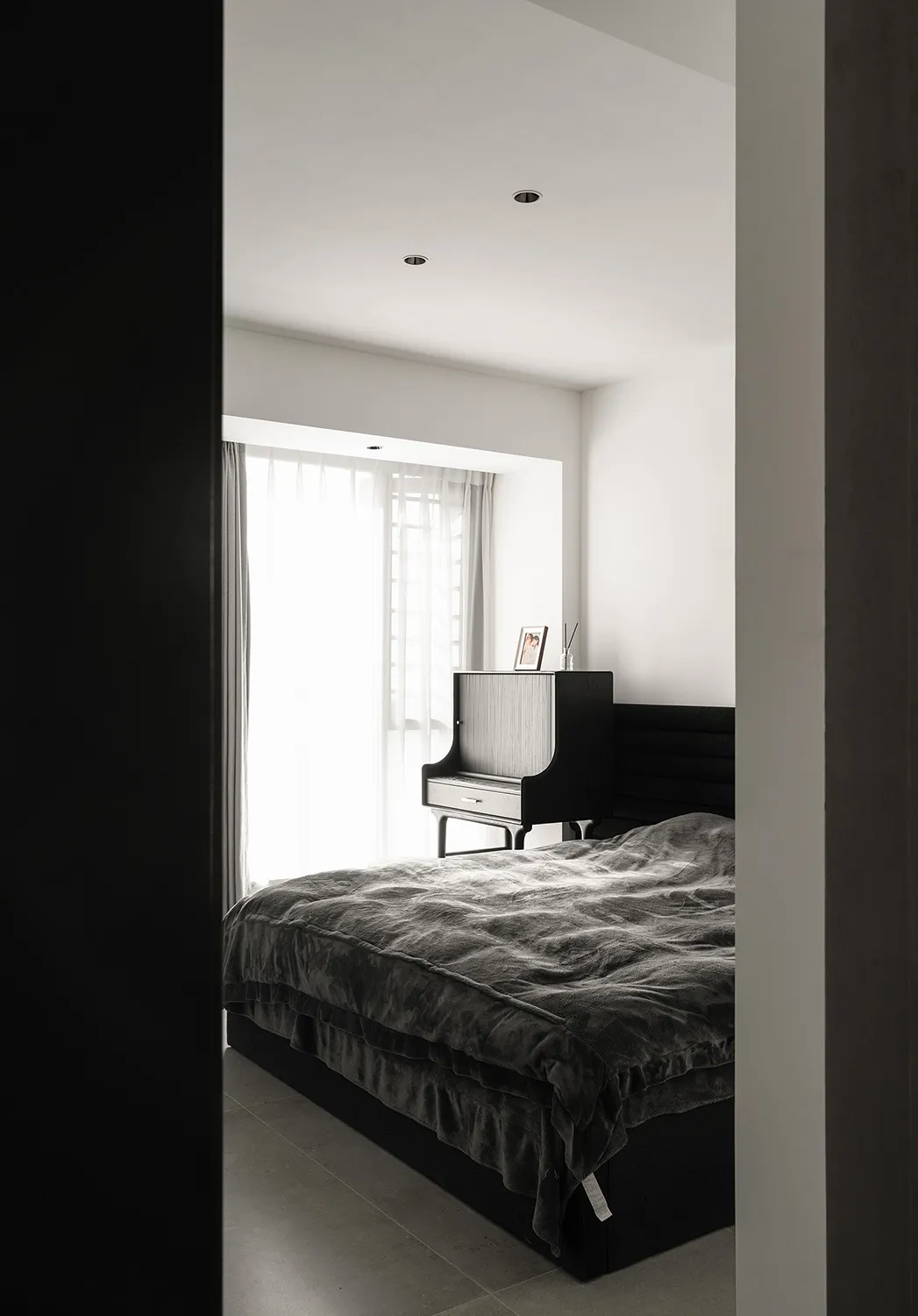 A modern bedroom with a black bed and grey blanket under bright sunlight from a large window.