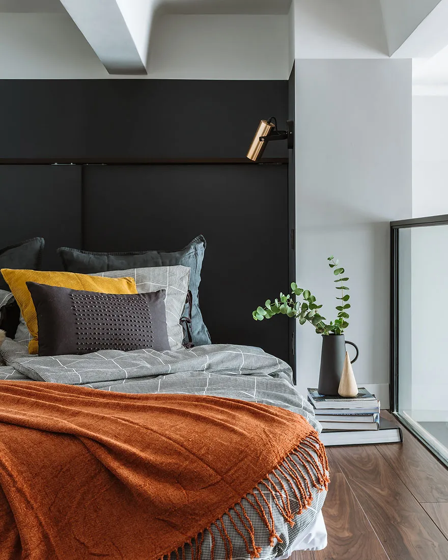 Minimalist bedroom featuring a bed with a dark headboard and mixed texture bedding, along with a small plant on a stack of bo