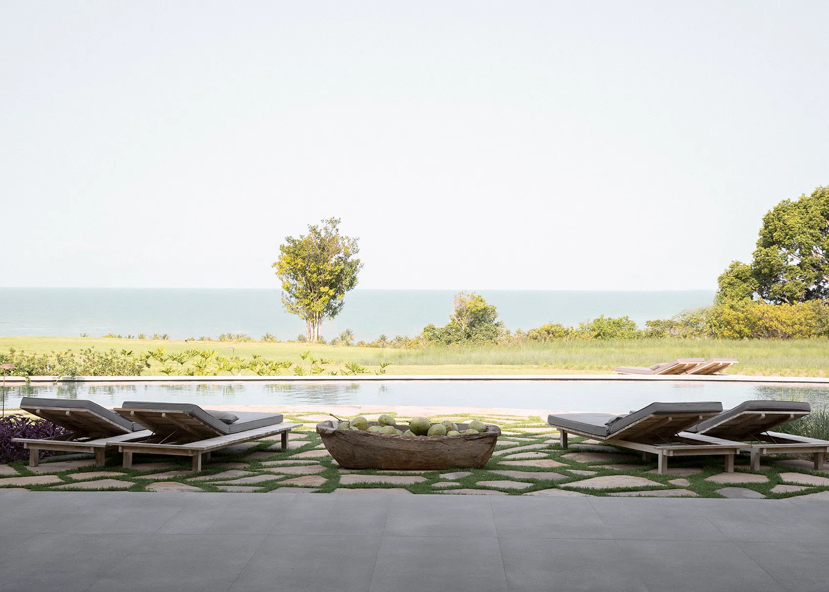 A minimalist residential patio featuring wooden furniture and a wooden bowl filled with green coconuts, overlooking a serene