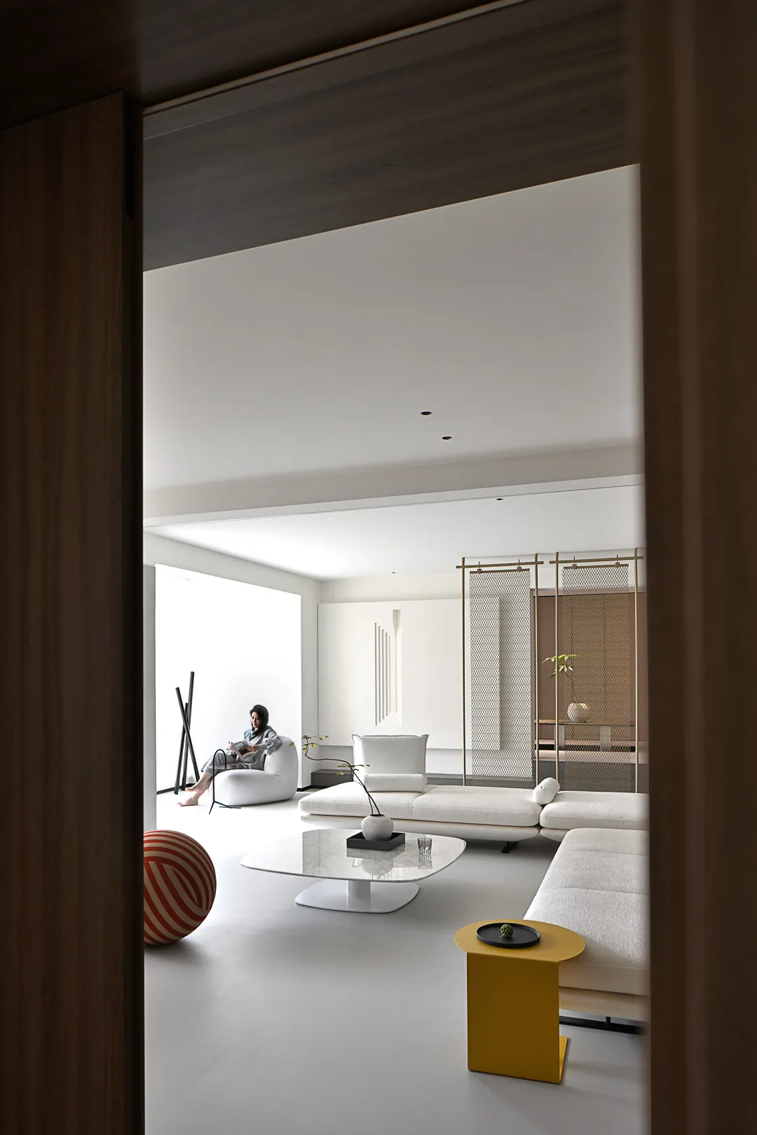 White minimalist living room with lounge sofas, a coffee table, and a round stool, featuring a woman sitting on a white sofa.