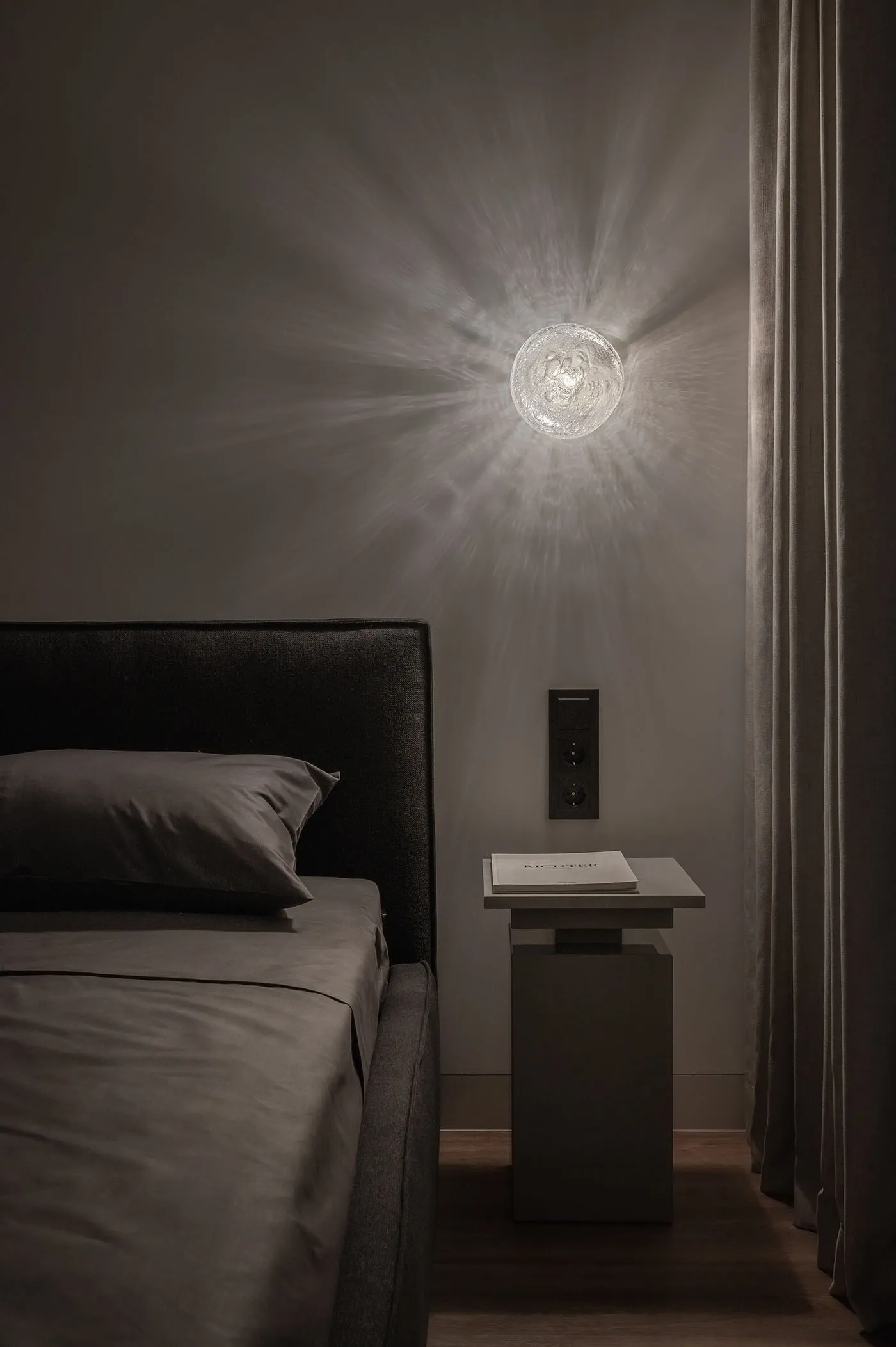 A modern bedroom featuring dark bedding and a wall-mounted light fixture casting a warm glow.