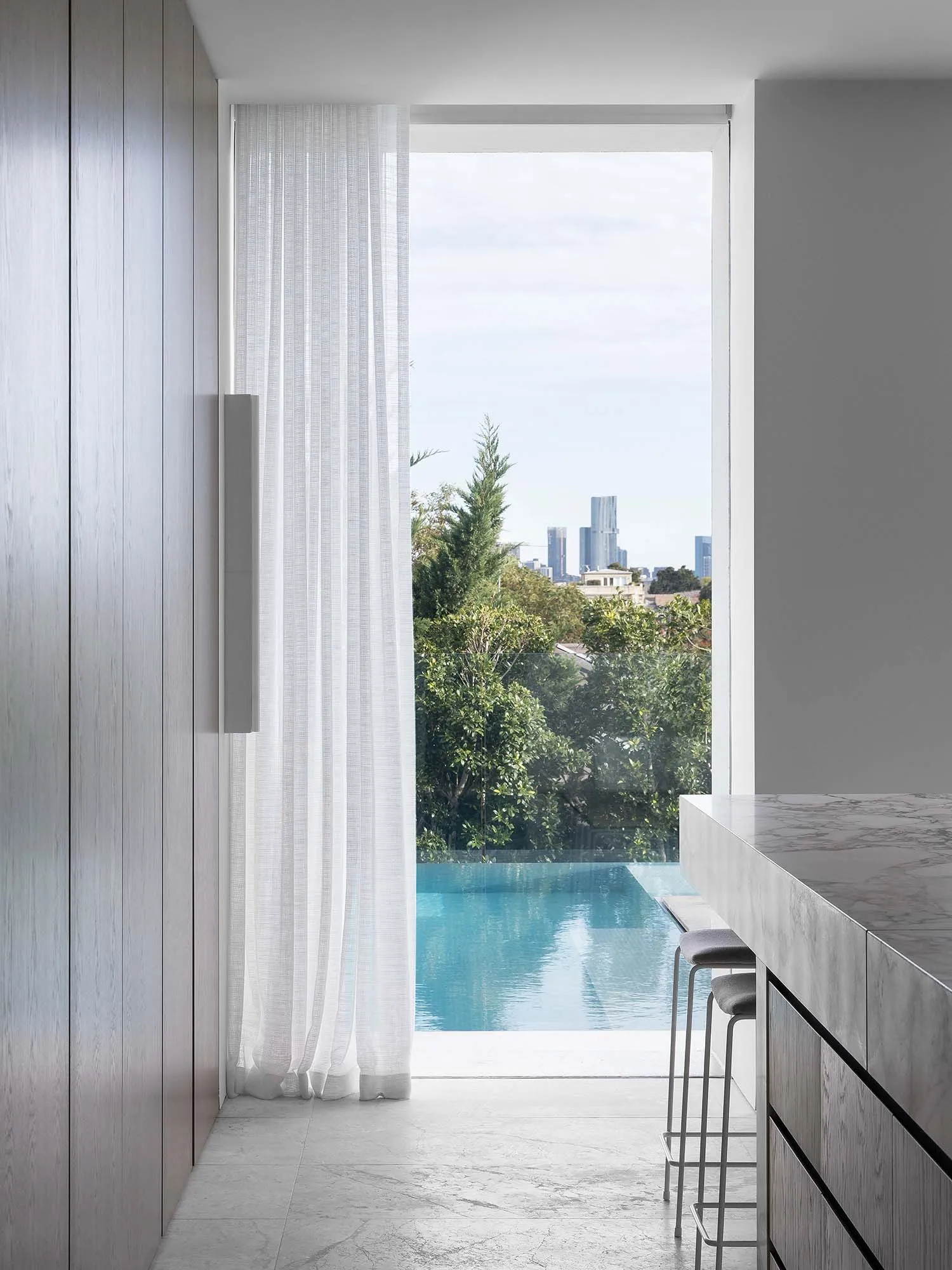 A modern residential kitchen features a glass window overlooking an infinity pool and city skyline, with marble countertops a
