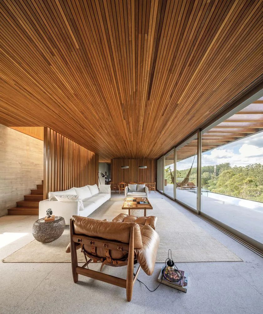 A modern living room featuring a wood-paneled ceiling and floor-to-ceiling glass windows, incorporating a mix of contemporary