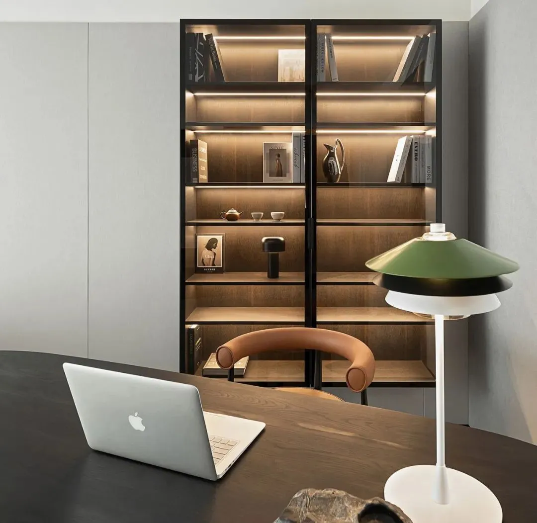 A modern minimalist office setup featuring a dark wooden desk, a green and black lamp, and a black bookshelf filled with book