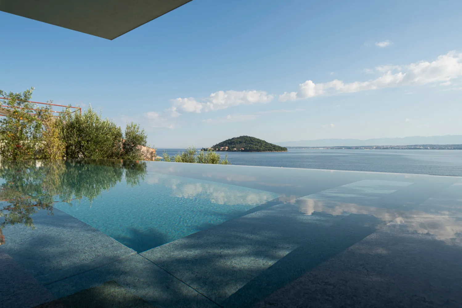 A minimalist swimming pool with a concrete deck and glass walls overlooks a calm sea.