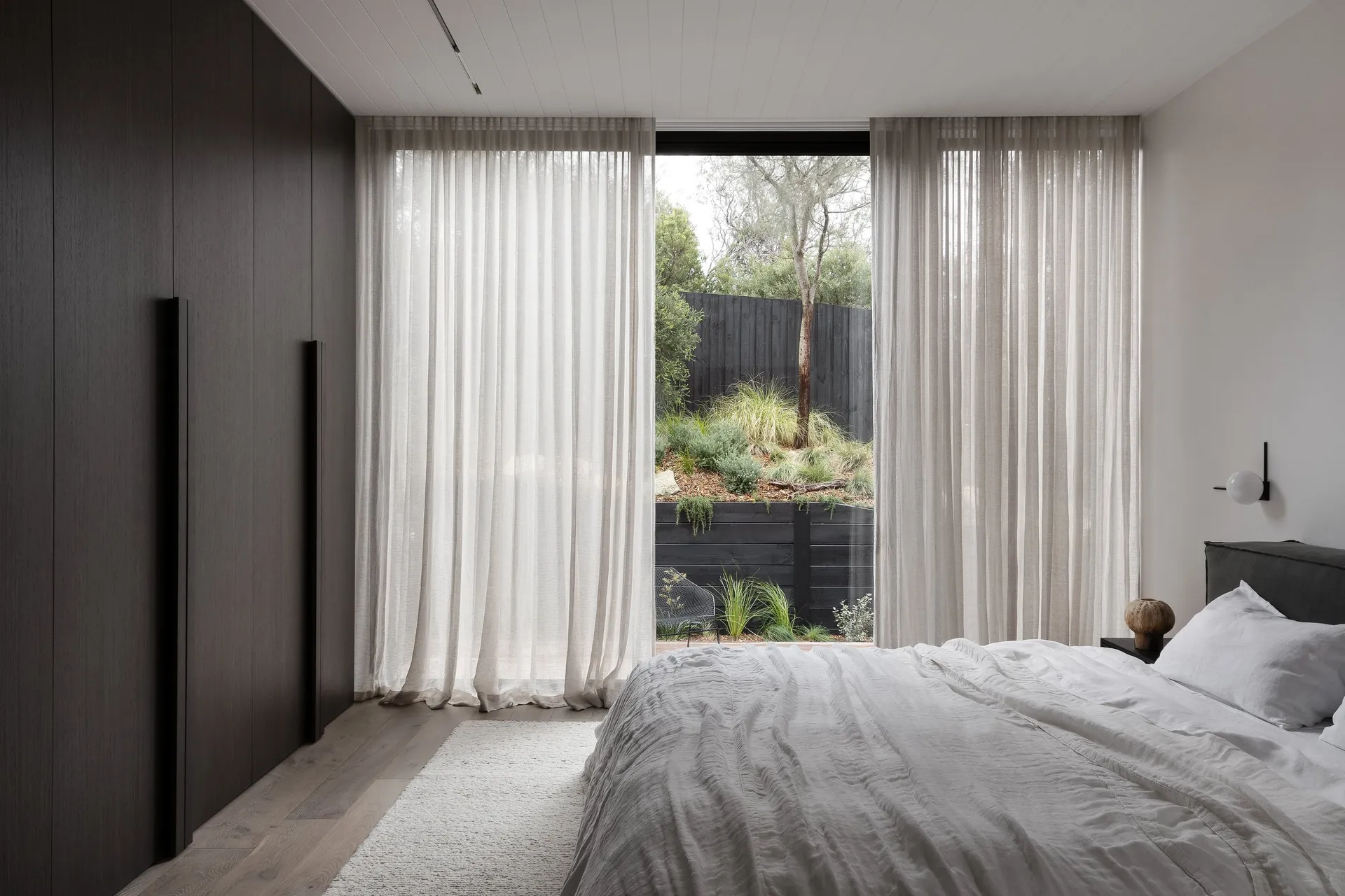 A minimalist bedroom featuring white curtains, a wooden floor, and a white bed with a view of greenery outside.