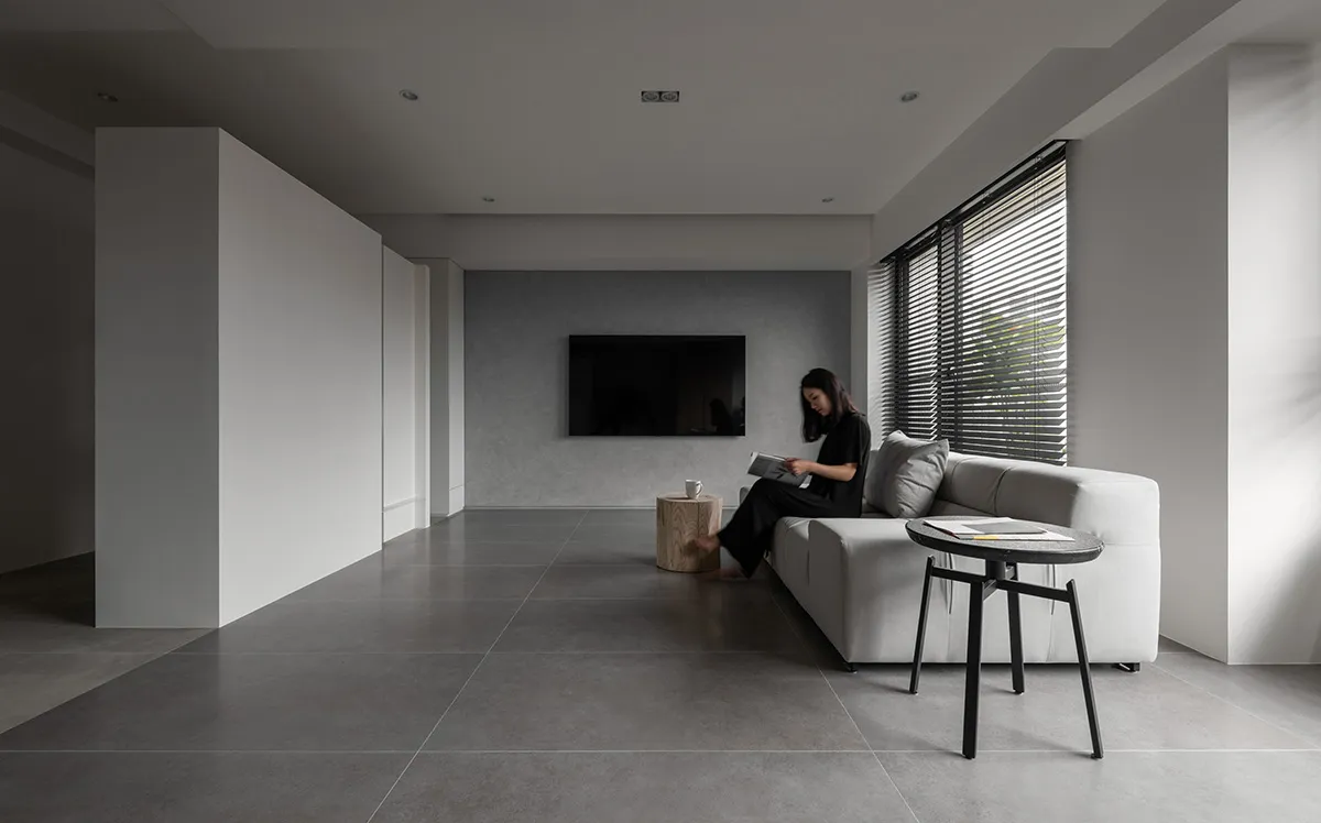 A modern minimalist living room with concrete flooring, a white sofa, and a reading woman sitting beside a window with blinds